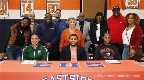 Group of people standing around three people that are seated at a long table. "EHS" is printed on the orange table cloth.